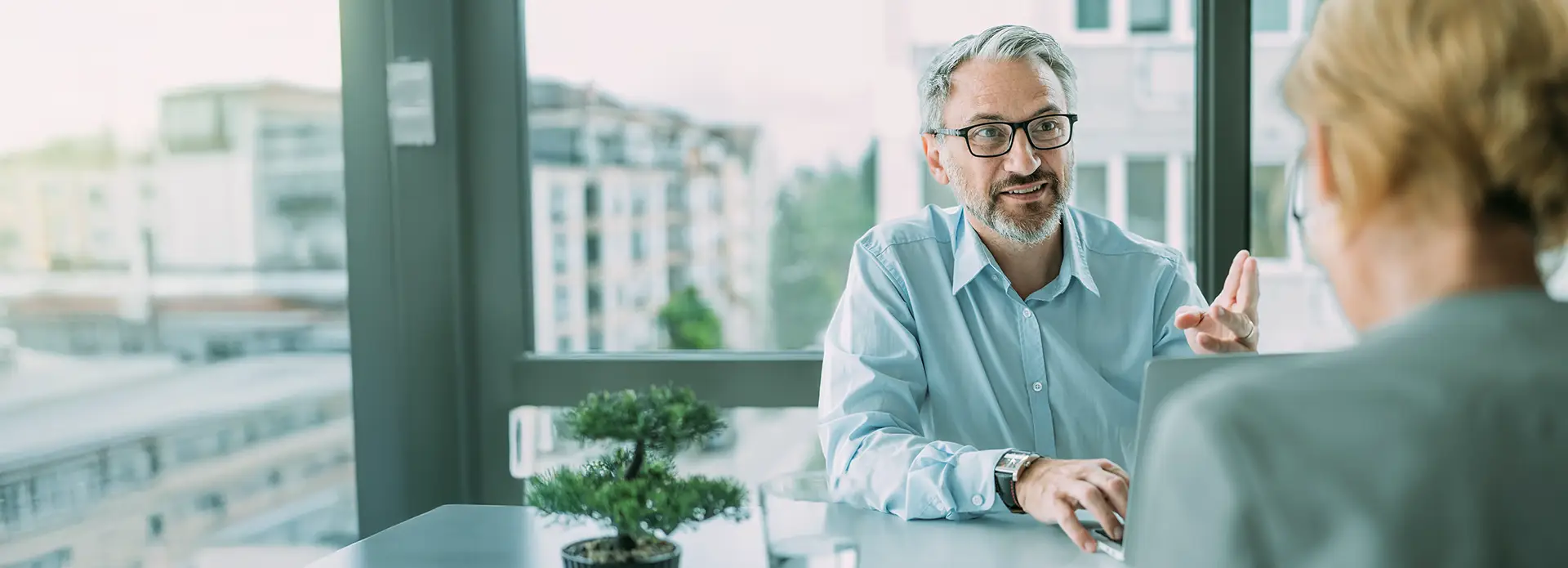Zwei Personen im Gespräch an einem Schreibtisch mit Laptop und Bonsai-Pflanze, im Hintergrund moderne Stadtarchitektur