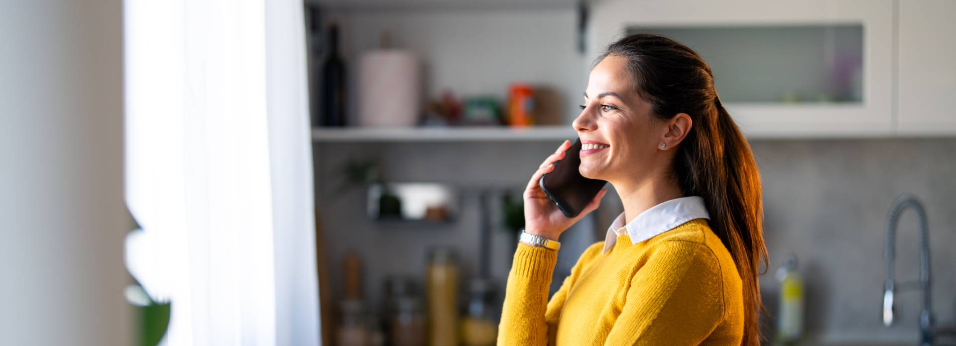 Frau in gelbem Pullover steht in einer modernen Küche und telefoniert lächelnd mit einem Smartphone.