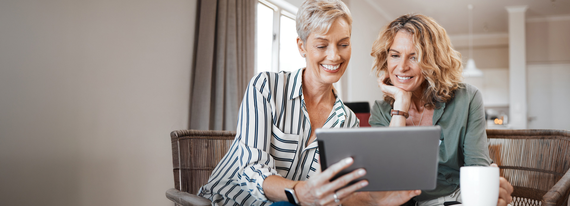 Zwei Frauen mittleren Alters sitzen in einem Wohnzimmer und schauen gemeinsam lächelnd auf ein Tablet.
